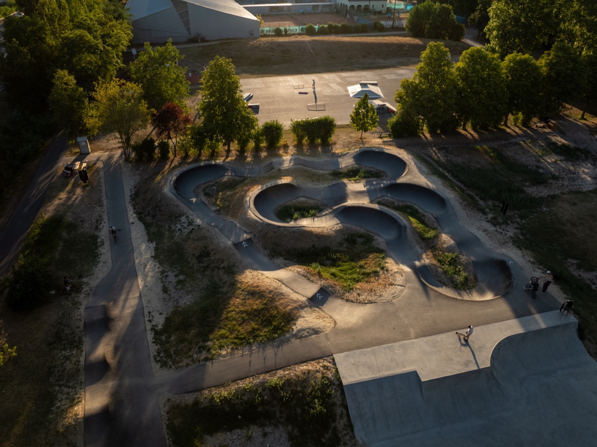 Luftaufnahme eines neuen Pump Tracks mit kleinen Fahrer:innen, im Hintergrund alter Bereich und Tennisplätze, Abendlicht.