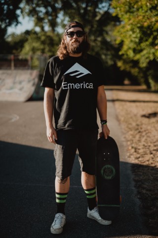 Karl Klang steht mit einem Skateboard vor einer Rampe am Schwarzen Platz, trägt schwarzes Emerica-Shirt und Sonnenbrille.