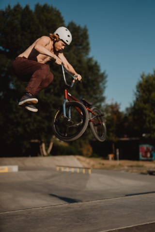 Max Gabriel führt am Schwarzen Platz einen Tailwhip auf seinem BMX vor, Skatepark im Hintergrund.