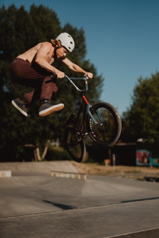 Person mit Helm springt mit BMX-Rad und verdrehtem Rahmen über eine Betonrampe, Bäume und blauer Himmel im Hintergrund