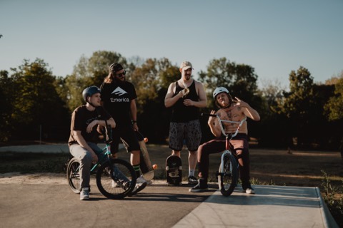 Vier Männer mit BMX-Rädern und Skateboards auf einer Skatepark-Rampe im Abendlicht, Bäume im Hintergrund.