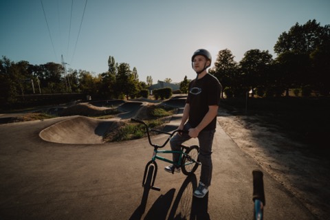 Ein Mann mit Helm steht mit BMX-Rad auf einer Bahn mit geschwungenen Erdmulden im Gegenlicht der Abendsonne.