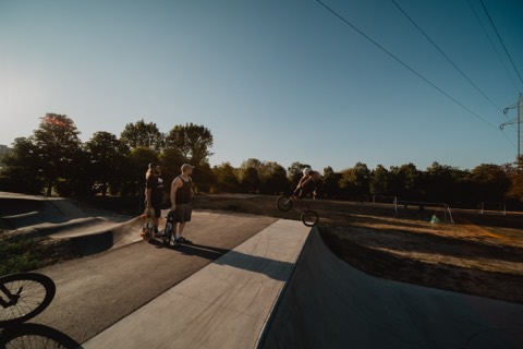 BMX-Fahrer springt mit gedrehtem Rad von einer Rampe, zwei Personen mit Skateboards schauen zu, Abendlicht.