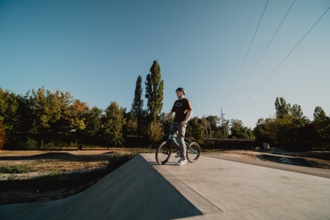 Person mit Helm steht mit BMX-Rad auf einer Betonrampe im Skatepark, Bäume und Freileitungen im Hintergrund.