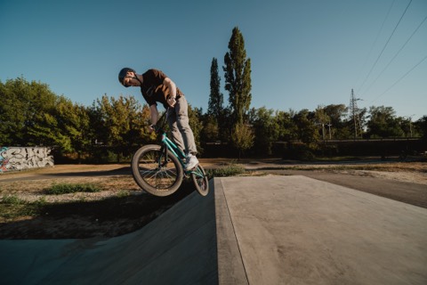 BMX-Fahrer mit Helm fährt auf einer Betonrampe im Skatepark, Bäume und Graffiti-Wand im Hintergrund