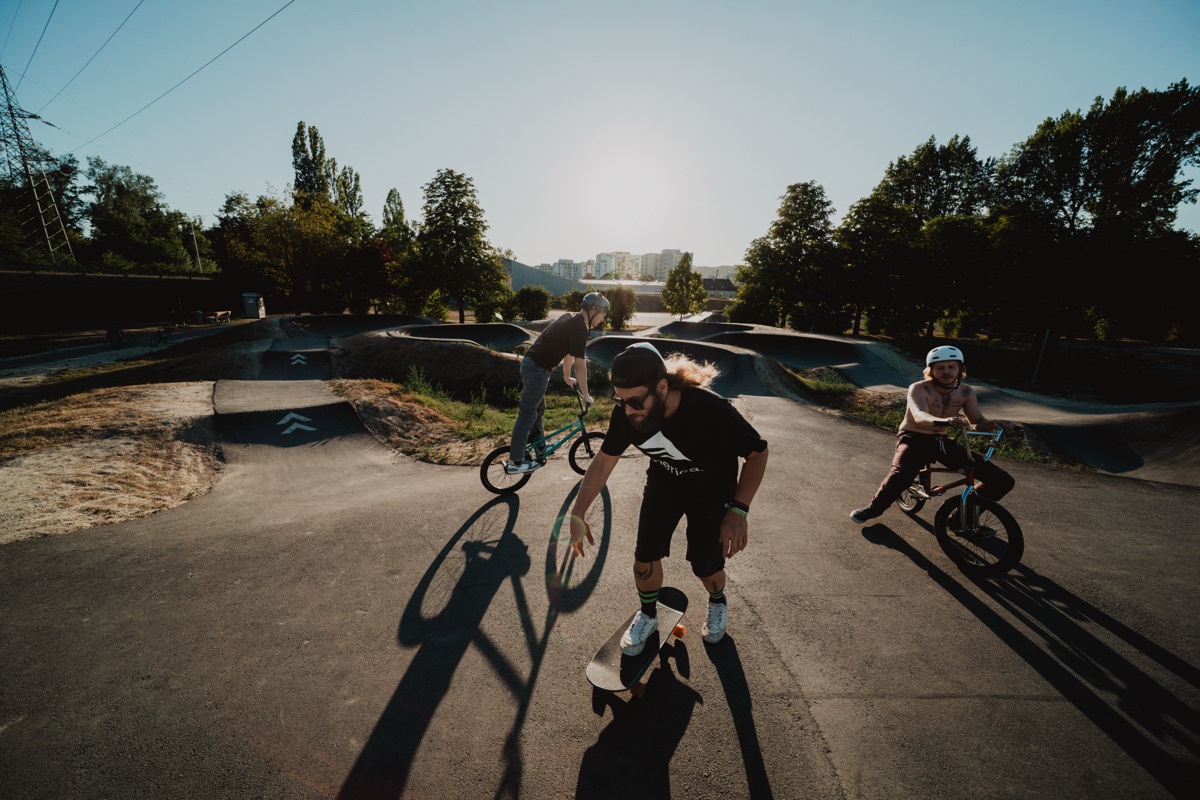 Drei Fahrer – ein Skateboarder und zwei BMX-Fahrer – starten auf einem Pumptrack in unterschiedliche Richtungen, lange Schatten im Gegenlicht.