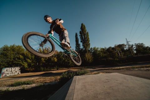 BMX-Fahrer springt mit gedrehtem Rad über eine Betonrampe, Graffiti-Wand und Bäume im Hintergrund.