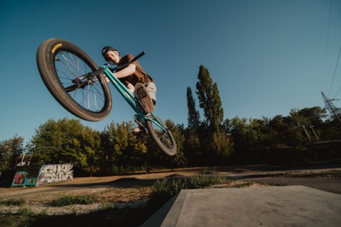 BMX-Fahrer springt mit gedrehter Lenkstange über eine Betonrampe, im Hintergrund Bäume und eine Graffiti-Wand.