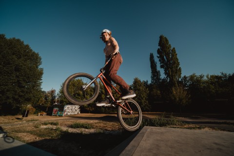 BMX-Fahrer mit Helm hebt das Vorderrad über eine Betonrampe, im Hintergrund Bäume und Abendhimmel.