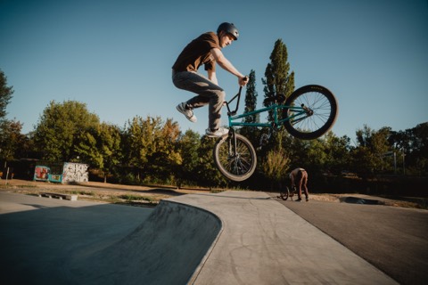 BMX-Fahrer Paul Leimer in der Luft mit seitlich gedrehtem Rad über einer Rampe, im Hintergrund eine Person mit Fahrrad.
