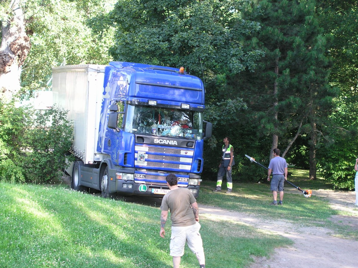 Ein blauer Sattelzug steht zwischen den Bäumen am Rand des Platzes, jemand schneidet mit einer Stangensäge Äste weg, damit er durchkommt