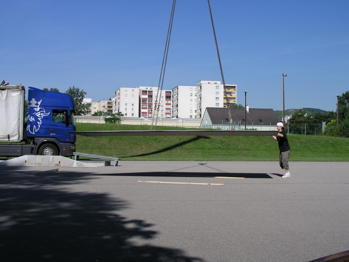 Ein Kran hebt ein langes flaches Rampenelement vom Lastwagen, eine Person führt es am unteren Ende