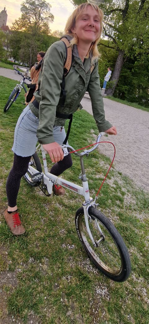 Lächelnde Frau auf weißem Fahrrad auf einer Wiese, dahinter eine weitere Person mit Fahrrad und ein Turm zwischen Bäumen.
