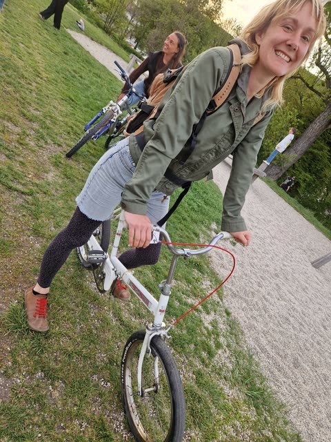 Lisi von der Black Place Society lächelt auf einem Fahrrad, im Hintergrund eine weitere Person auf einem Rad im Grünen